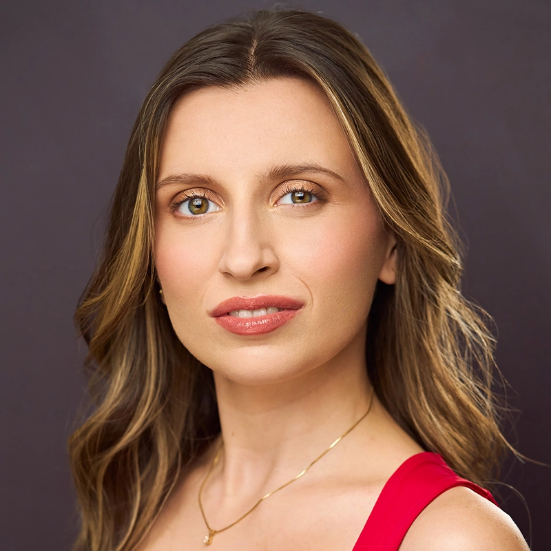 A woman with wavy brown hair and hazel eyes wears a sleeveless red top and a thin gold necklace. She has natural makeup and a neutral expression. The background is a smooth dark gray, giving the portrait of Jillian Millette an understated elegance.