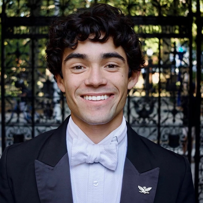 A young man with curly dark hair, wearing a tuxedo and white bow tie, smiles at the camera. A metal pin is on his left lapel. Behind him is an ornate iron gate with sunlight filtering through trees. This elegant portrait captures Benjamin Jimenez’s poised and cheerful presence against the picturesque backdrop.