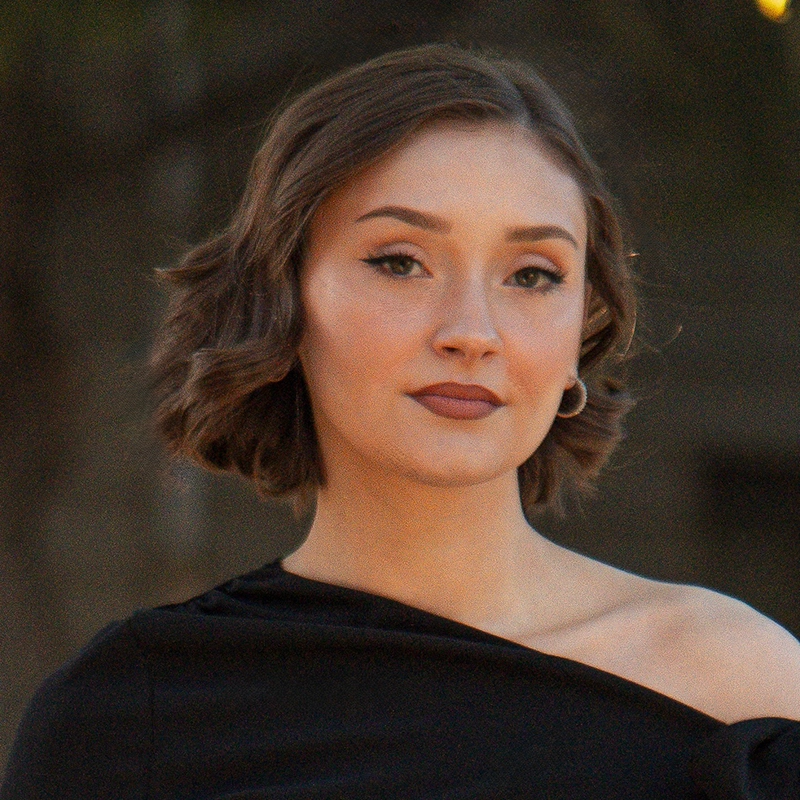 A woman with short wavy brown hair and hoop earrings wears an off-shoulder black top. She has winged eyeliner, bold lipstick, and a neutral expression. The background is blurred with hints of greenery and warm light.