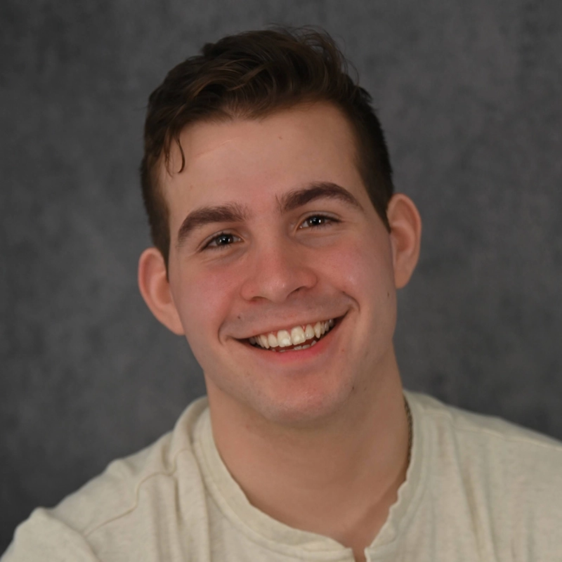 Jonathan Uber, a young man with short brown hair, smiles warmly at the camera. He is wearing a light beige, buttoned shirt and is posed in front of a dark, textured gray background. The soft lighting highlights Jonathan’s face and creates a friendly atmosphere.