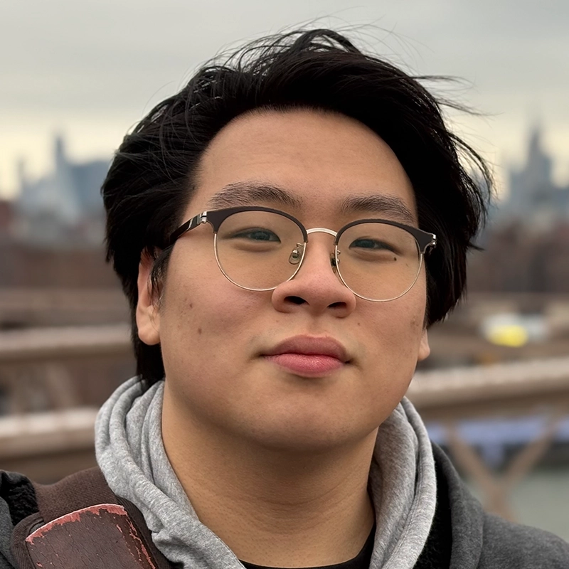 A young person with short black hair and round glasses stands outdoors, wearing a gray hoodie and dark jacket. The background is blurred, showing an urban skyline under a cloudy sky. The person looks directly at the camera with a neutral expression.