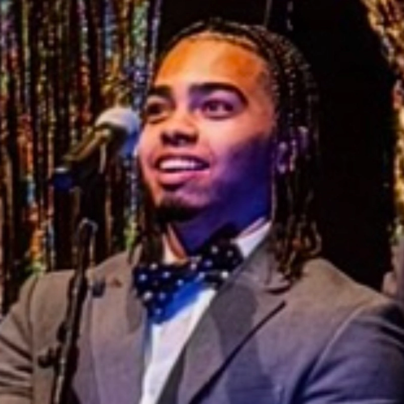 Josue Tejada with braided hair smiles while speaking into a microphone, wearing a grey suit jacket and a dark bow tie with light-colored dots.