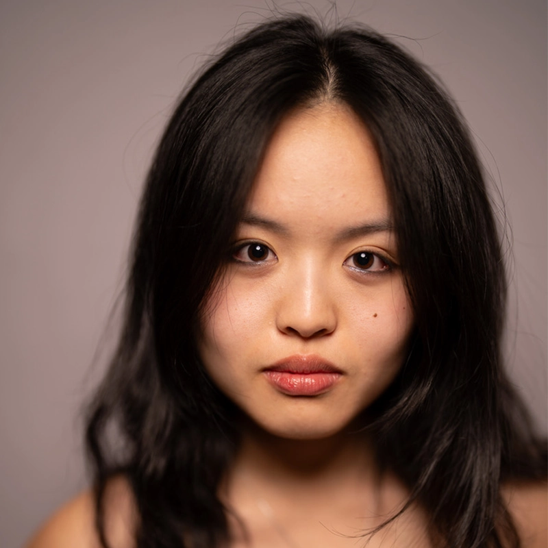 A young woman with long black hair and smooth skin stares directly at the camera. She has full lips, dark eyes, and a neutral expression. The background is blurred and light gray, emphasizing her face—capturing the quiet confidence of Julia Hayden Fung.