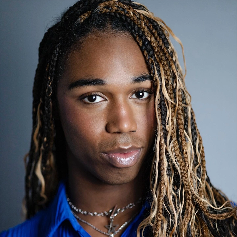 Phoebe Kleehammer, with long, highlighted braids, gazes at the camera while wearing a blue shirt and layered silver necklaces. Their makeup is subtle, featuring glossy lips and soft eyeshadow. The background is a neutral gray, keeping focus on their expressive face.