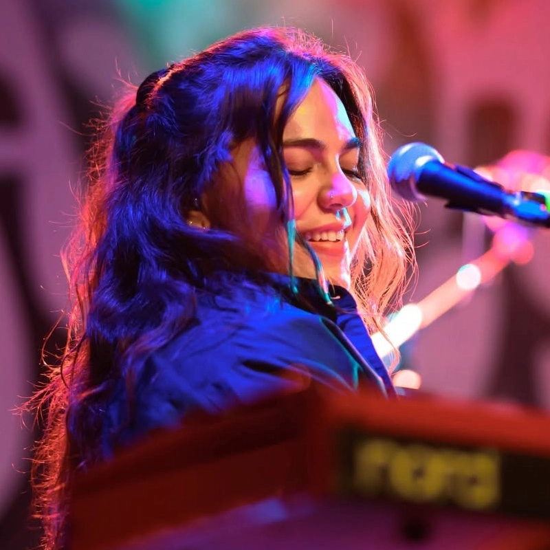 A woman with long wavy hair smiles while singing into a microphone. She stands behind a red Nord keyboard, under colorful stage lighting with pink, blue, and orange hues. The background is blurred with abstract shapes.