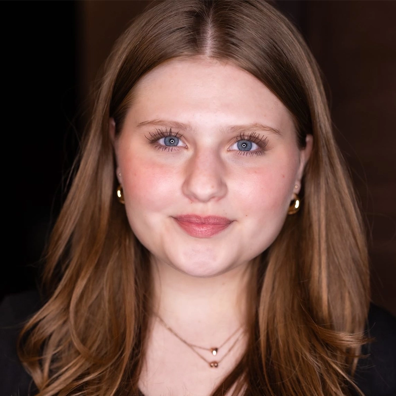 Phoebe Kleehammer, a young woman with light skin, long light-brown hair, blue eyes, and pink lipstick, smiles slightly. She wears gold hoop earrings, layered necklaces, and a black top. The background is dark and out of focus.