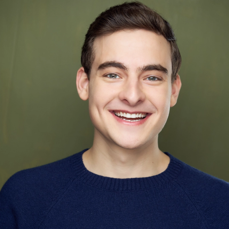 A young man with fair skin, short brown hair, and blue eyes smiles broadly. He wears a dark blue sweater. The background is a plain olive green. The image is brightly lit, highlighting his cheerful expression.