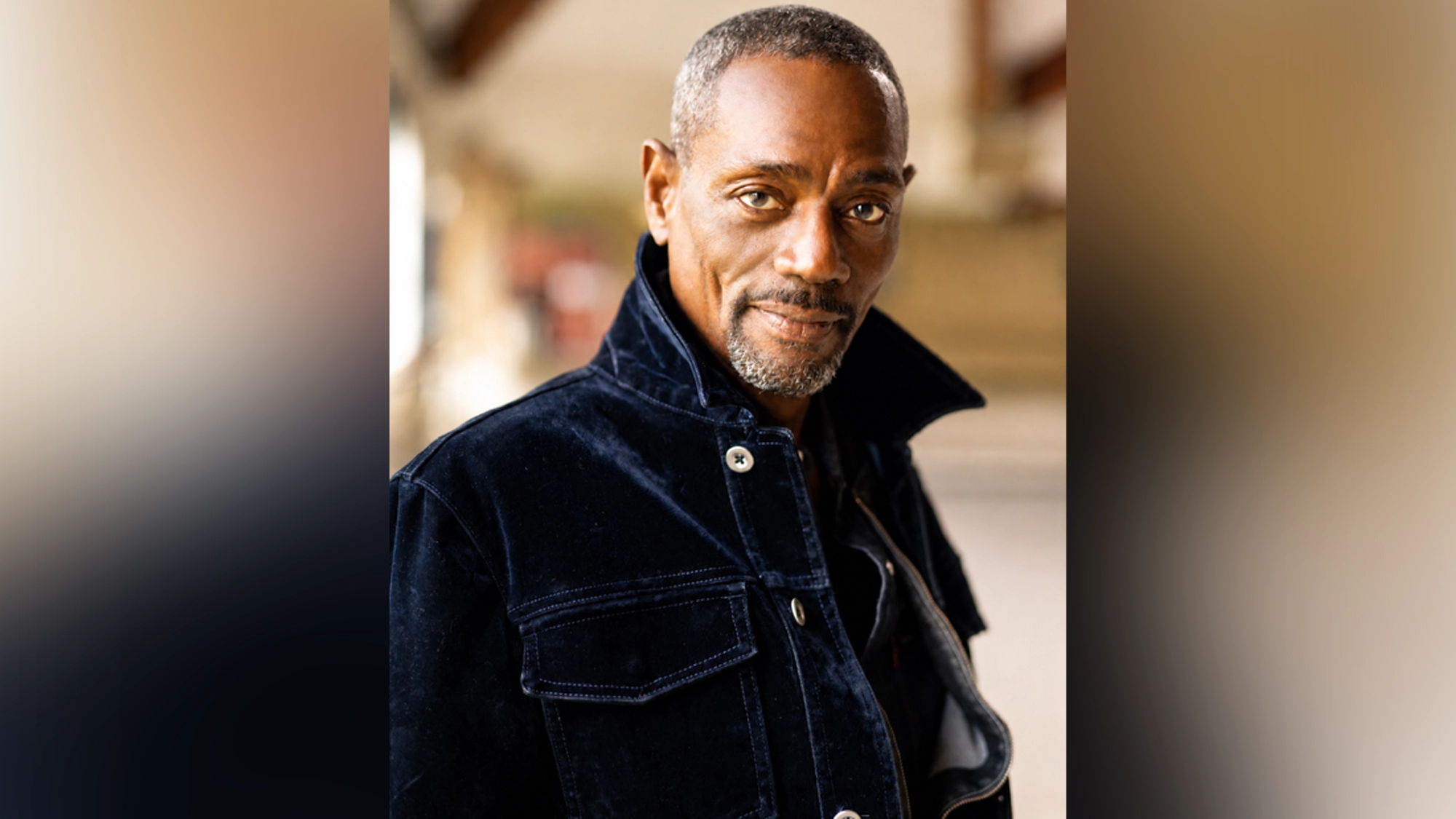 A middle-aged man with short graying hair and a goatee looks confidently at the camera. He wears a dark blue corduroy jacket with the collar popped, evoking the cool tones of blues ballads. The background is softly blurred, suggesting an outdoor urban setting.