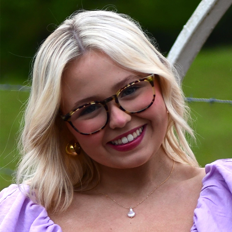 Kinsley Stephens, a young woman with wavy blonde hair, tortoiseshell glasses, and gold earrings, smiles outdoors. She wears a light purple top and a pearl necklace. A blurred green background and a wire fence are visible behind her.