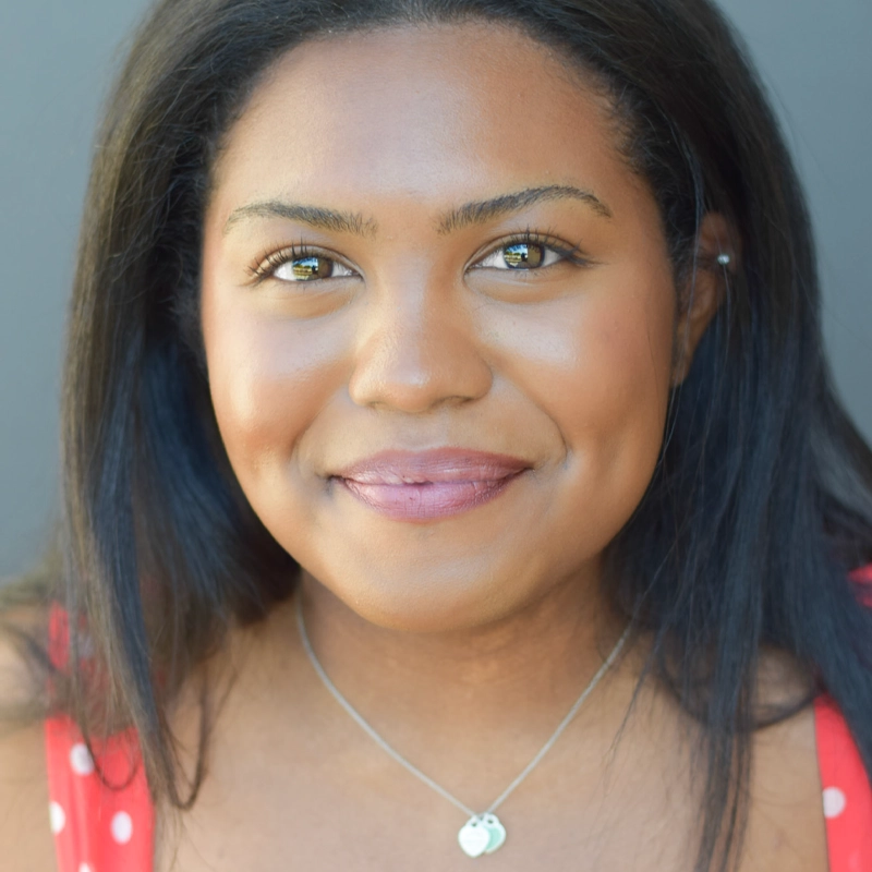A woman with medium-dark skin, straight dark hair, and brown eyes smiles softly. She wears a red polka dot top, a small silver heart necklace, and natural makeup. The background is plain and out of focus, allowing the warmth of her expression—reminiscent of Kaleo Knight’s gentle charm—to stand out beautifully.