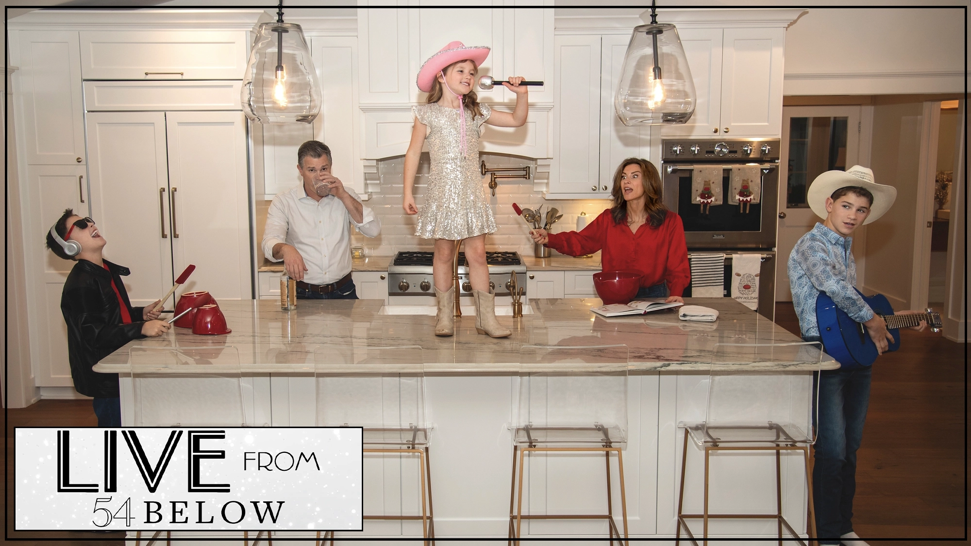 A family poses in a kitchen; a girl in a sparkly dress and pink hat sings on the island, a boy in a cowboy hat plays guitar, a man eats, a woman stirs batter, and another boy wears sunglasses and sings. “LIVE from 54 BELOW” is written in the corner, bringing to mind an energetic livestream performance featuring Jenny Powers and Matt Cavenaugh.