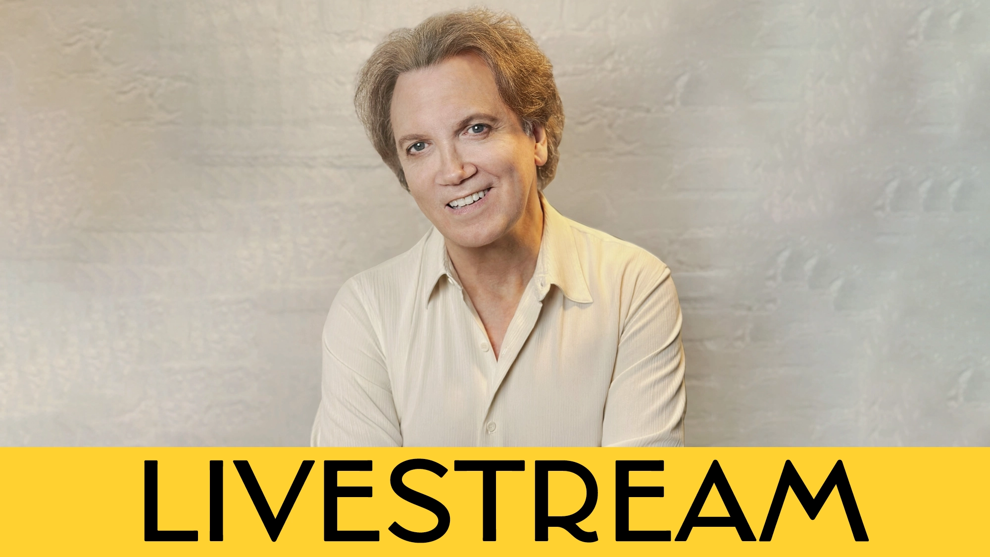 A smiling person with short, styled light brown hair, wearing a beige shirt, poses in front of a light textured wall. Across the bottom, a yellow bar displays the bold black text: “LIVESTREAM,” highlighting a special event featuring Charles Busch and classic tunes by Johnny Mercer.