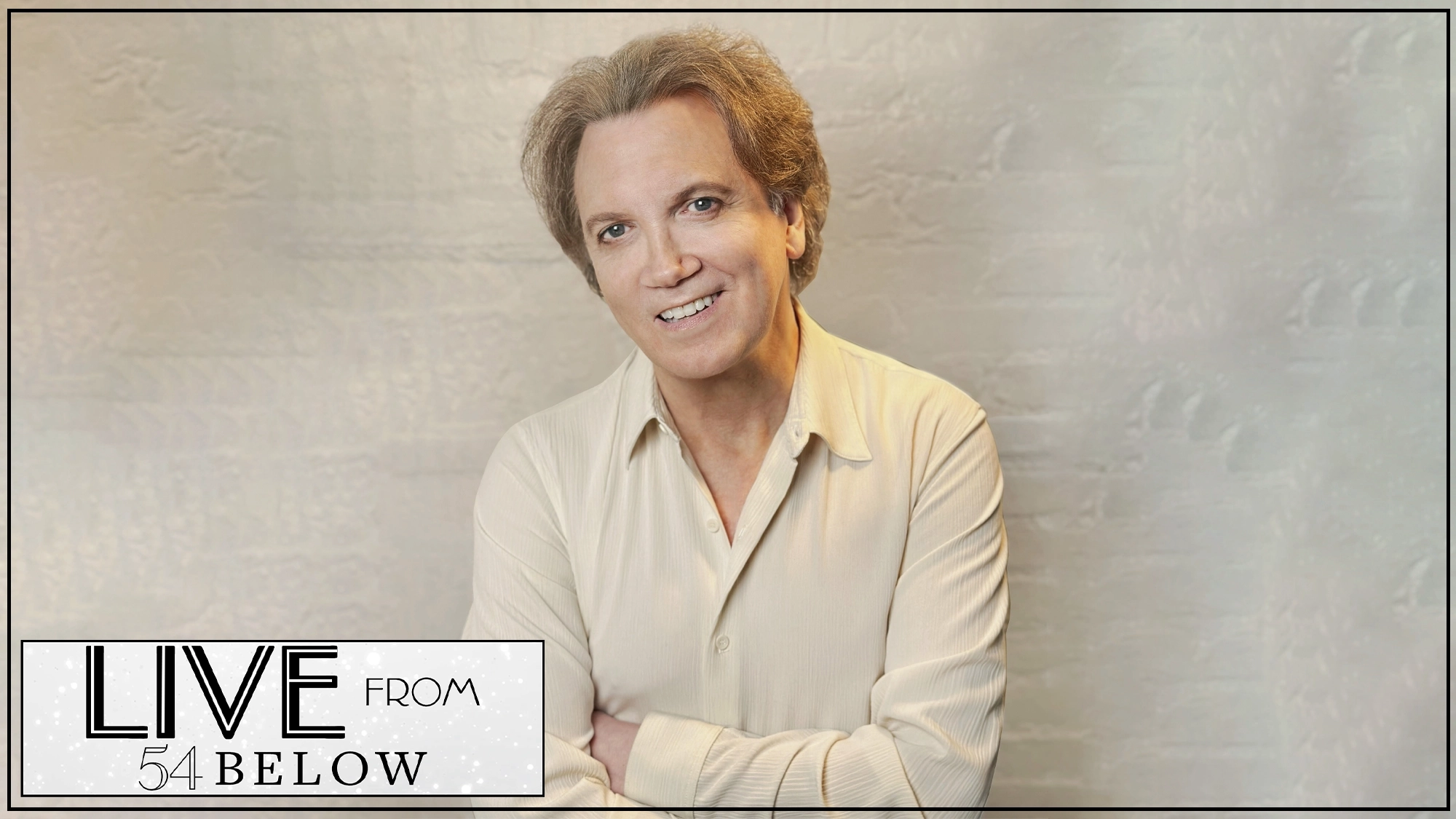 A smiling man with light brown hair, wearing a cream-colored button-up shirt, stands against a pale textured background. His arms are loosely crossed. In the bottom left corner, a graphic reads “LIVE FROM 54 BELOW,” hinting at an upcoming livestream event celebrating talents such as Charles Busch and Johnny Mercer.