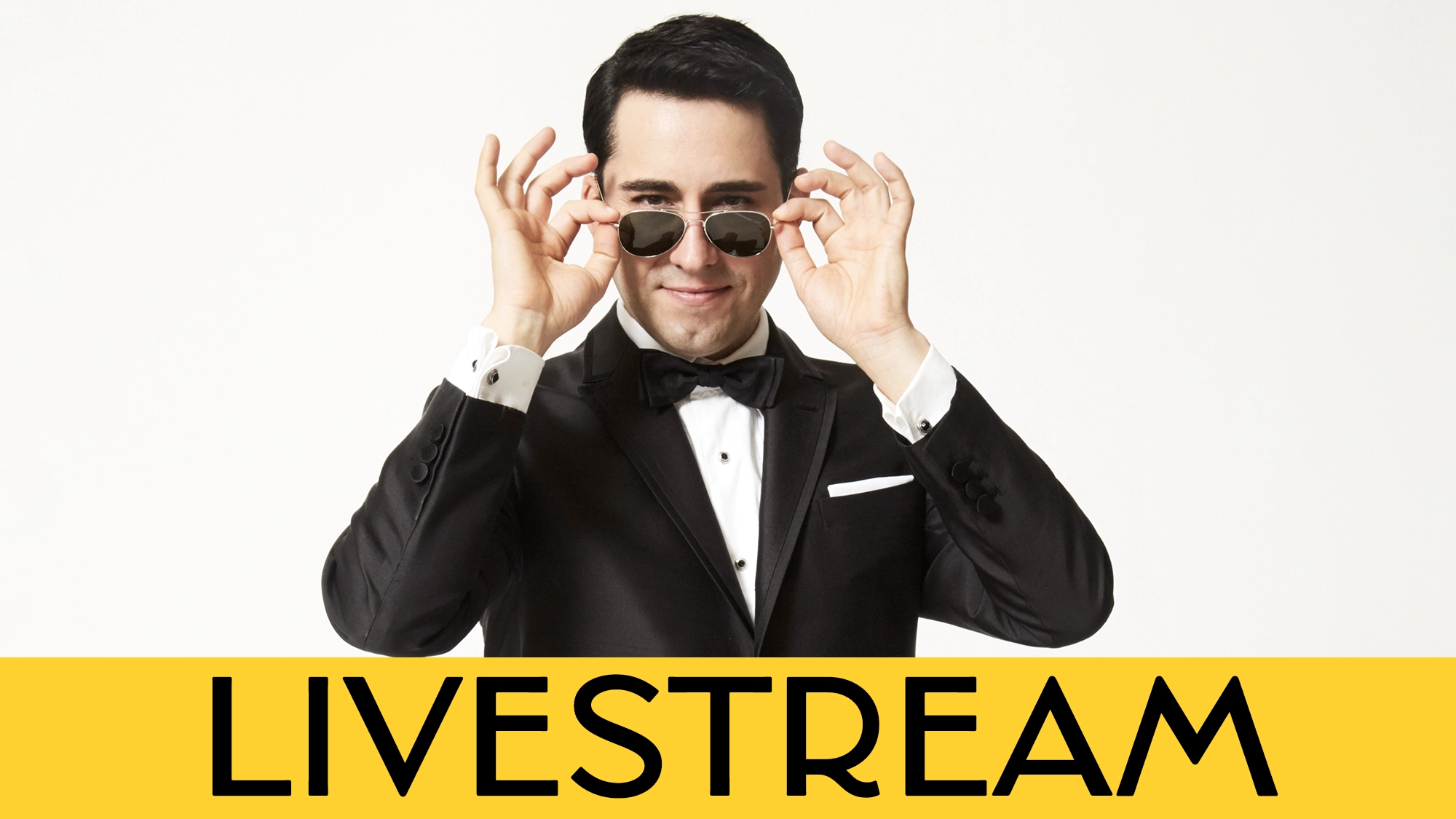 LIVE JLY Calendar A man in a tuxedo lifts sunglasses while smiling against a white background. The lower part of the image features a yellow banner with the word “LIVESTREAM” in bold black letters, highlighting John Lloyd Young’s Songs From the Road Livestream.