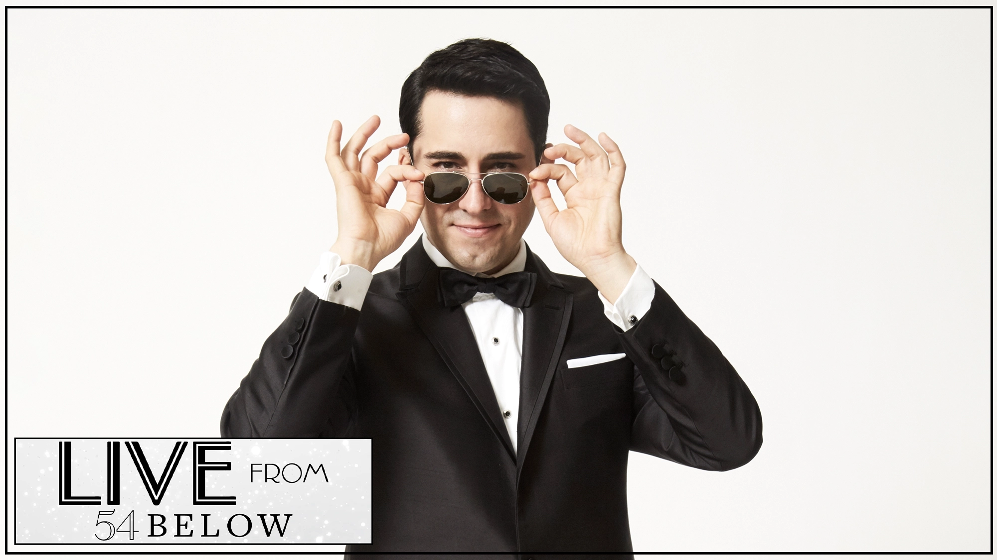 A man in a black tuxedo and bow tie stands against a white background, smiling as he lifts dark sunglasses. In the bottom left corner, there is a graphic that reads “LIVE FROM 54 BELOW” in bold, artistic text—capturing the excitement of John Lloyd Young’s Livestream performance of Songs From the Road.