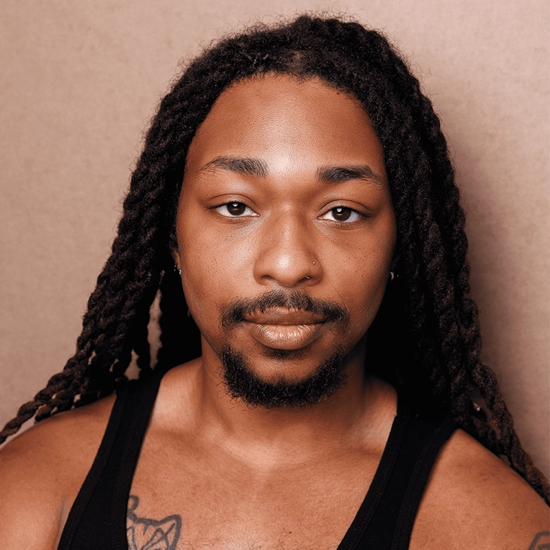 A person with long, dark, twisted hair, light brown skin, and a trimmed beard looks at the camera. Spencer Bowman wears a black tank top, revealing part of a chest tattoo, and has a nose piercing. The background is plain and neutral beige.
