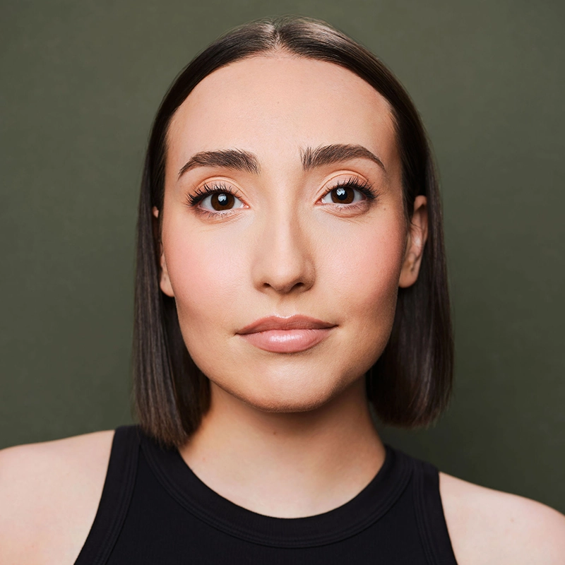 A young woman, Leah Mossman, with straight, shoulder-length brown hair and full eyebrows looks directly at the camera. She wears a sleeveless black top and subtle makeup. The background is solid olive green. Her expression is neutral and composed.