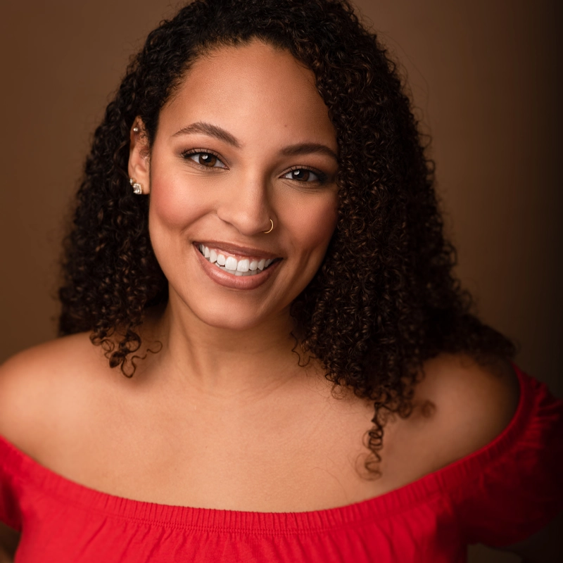 A smiling woman with curly hair wears a red off-the-shoulder top. She has a nose ring and stud earrings. The background is plain brown, and she is looking directly at the camera. The lighting is warm and soft, highlighting her features—capturing Siva Lissette’s vibrant personality in every detail.