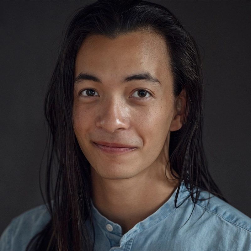 A person with long dark hair and light skin smiles softly at the camera. They wear a light blue, buttoned shirt and are set against a plain, dark background, creating a calm and natural portrait.