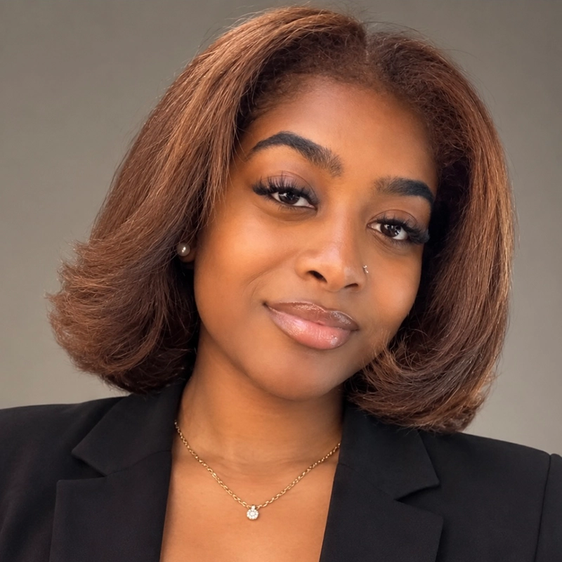 A young woman, Rayana D. Briggs, with medium brown shoulder-length hair styled in soft waves smiles gently. She wears a black blazer, a gold necklace with a small pendant, pearl earrings, and has a nose stud. The background is neutral gray and well-lit.