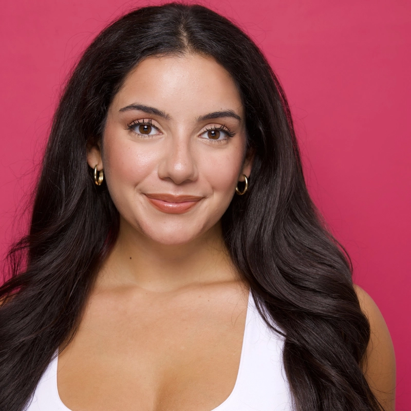 A woman with long, dark, wavy hair and gold hoop earrings smiles gently against a solid bright pink background. She wears a white top and natural makeup, giving a friendly, approachable appearance. Her hair is parted in the middle and cascades over her shoulders, perfectly capturing Jacqliene Mangini’s signature style.