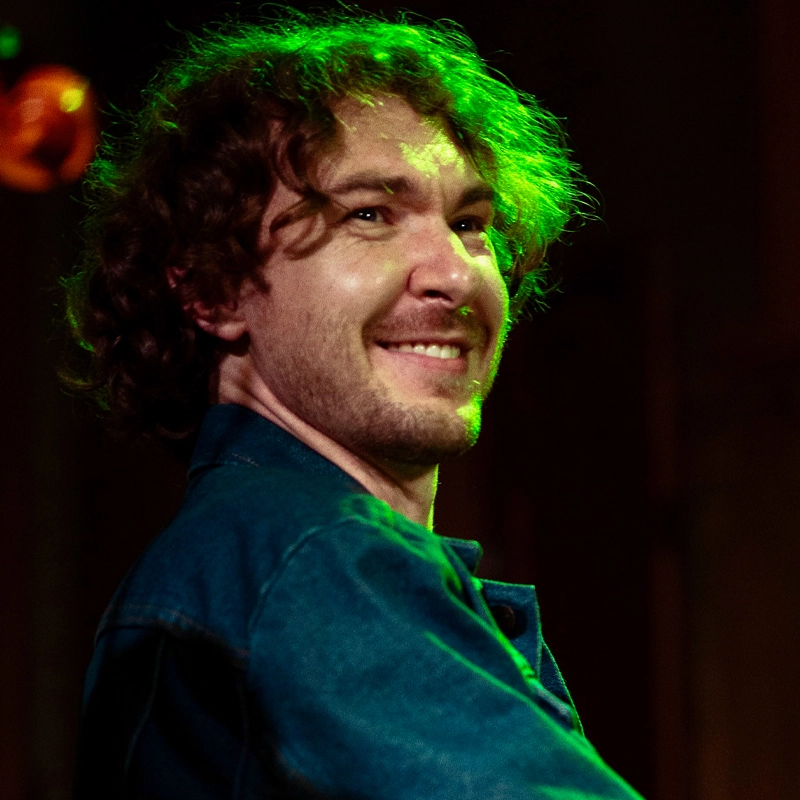 A smiling man, Roscoe McDonald, with curly brown hair stands under green stage lighting, wearing a denim jacket. The background is dark and out of focus, with an orange light fixture visible in the upper left corner. He appears to be on stage or at a performance.