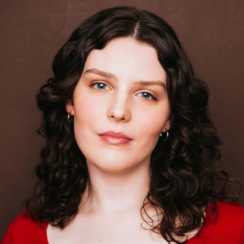 Melina Walko, a woman with fair skin, blue eyes, and shoulder-length wavy brown hair, wearing small hoop earrings and a red top, faces the camera with a neutral expression against a plain brown background.