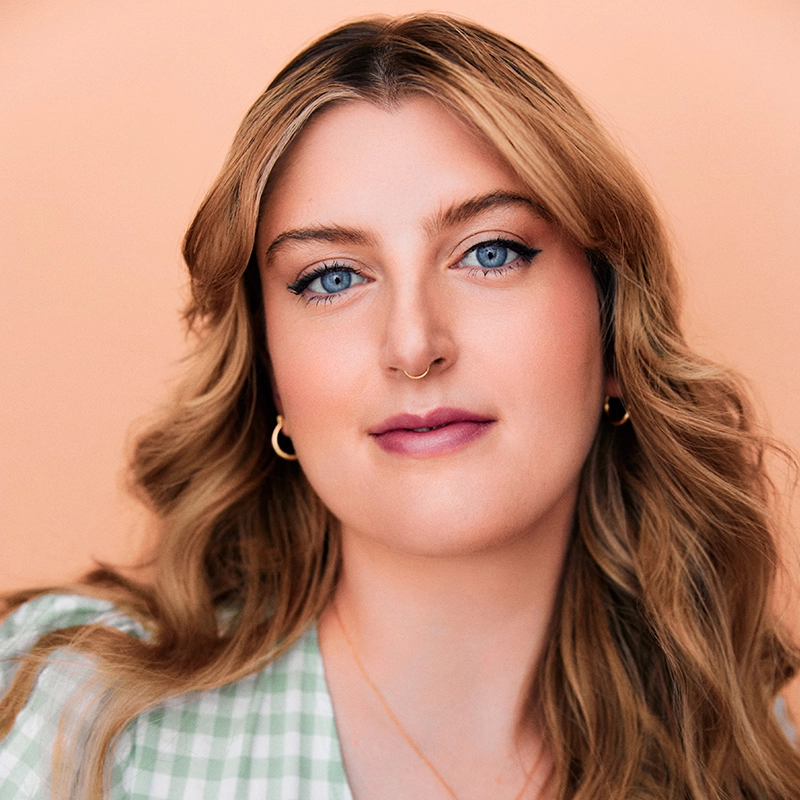 A woman with wavy blonde hair and blue eyes looks at the camera. She wears gold hoop earrings, a gold necklace, and a nose ring. Her makeup is subtle, and she wears a green gingham top. The background is peach-colored, highlighting Leah Mossman’s effortlessly chic style.
