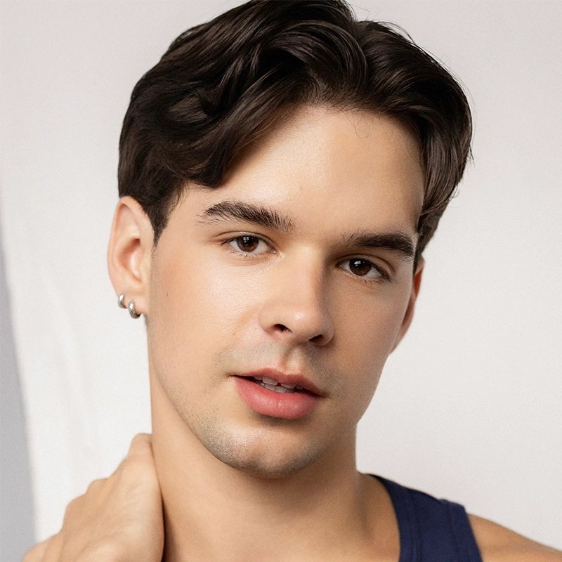 A young person with short, dark hair and smooth skin looks at the camera. They wear a dark tank top and a small hoop earring, gently touching their neck, with a neutral facial expression against a simple, light background—a style reminiscent of Leah Mossman’s modern portraiture.