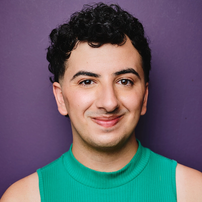 Spencer Bowman, a person with short, curly dark hair, smiles gently at the camera. They wear a sleeveless green top and stand against a plain purple background. Their expression is friendly and approachable.