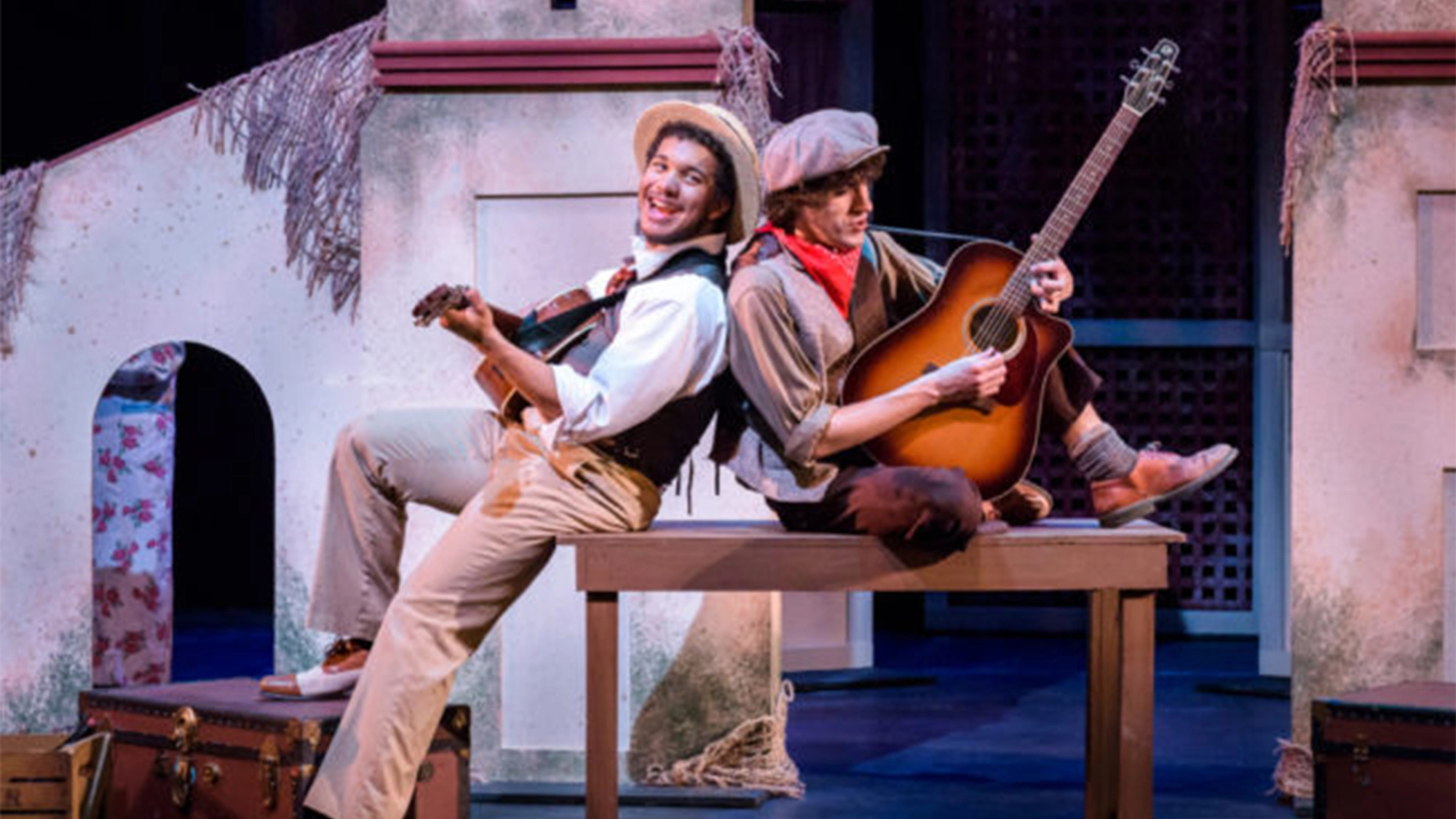 Two actors, Da’Von T. Moody and Cole Larravide, in period costumes sit back-to-back, each playing acoustic guitars. One sits on a table, the other on a suitcase. They smile joyfully. The stage set includes rustic, weathered structures, suggesting an outdoor or village scene.