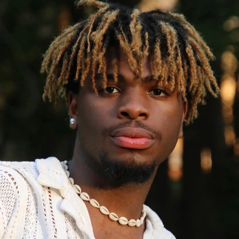 A young man with light brown and blonde dreadlocks stands outdoors, wearing a white shirt, shell necklace, and pearl earrings. He looks slightly off-camera with a calm expression. Sunlight softly illuminates his face, and greenery is blurred in the background—a serene moment reminiscent of a Gabriela Descartes López portrait.