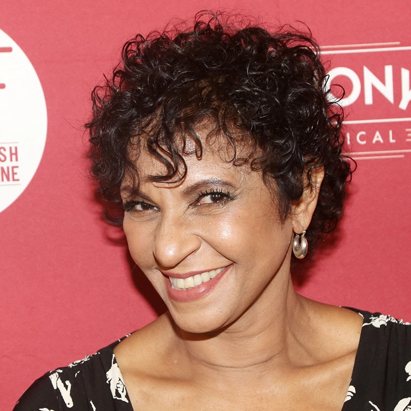 Nancy Ticotin, a woman with short curly black hair, wearing silver hoop earrings and a black-and-white patterned top, smiles at the camera in front of a red backdrop with white text and logos.