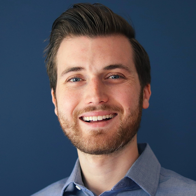 Neil McNulty, a man with fair skin, short brown hair, and a trimmed beard, smiles at the camera. He wears a light blue collared shirt and stands against a solid dark blue background. The lighting is even, highlighting his face and friendly expression.