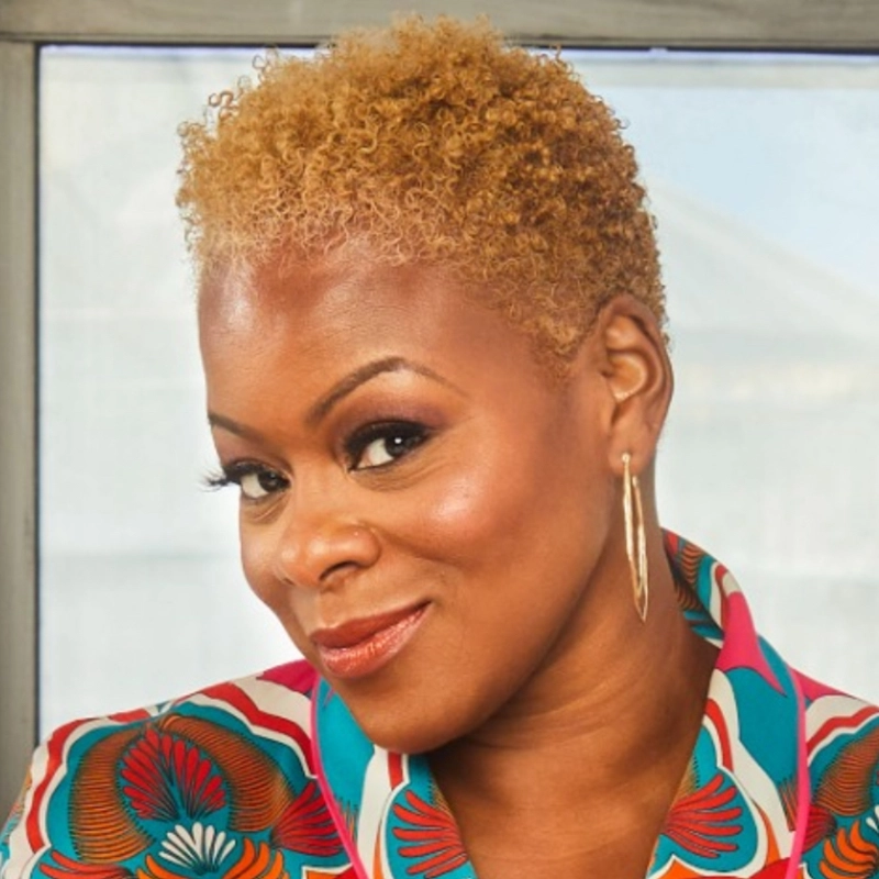 A woman with short, curly blond hair smiles slightly. She wears gold hoop earrings and a colorful, patterned top with red, blue, and white designs. The background is soft-focus, featuring a window and bright outdoor light. The confident energy in her expression suggests she could be Nikki Kimbrough.