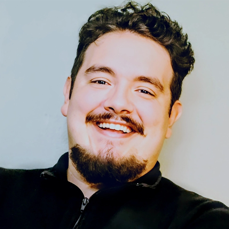 A young man with curly dark hair, a trimmed goatee, and a mustache smiles brightly. He is wearing a black zip-up top and is posed against a plain light-colored background, facing slightly left. The lighting is bright and even.