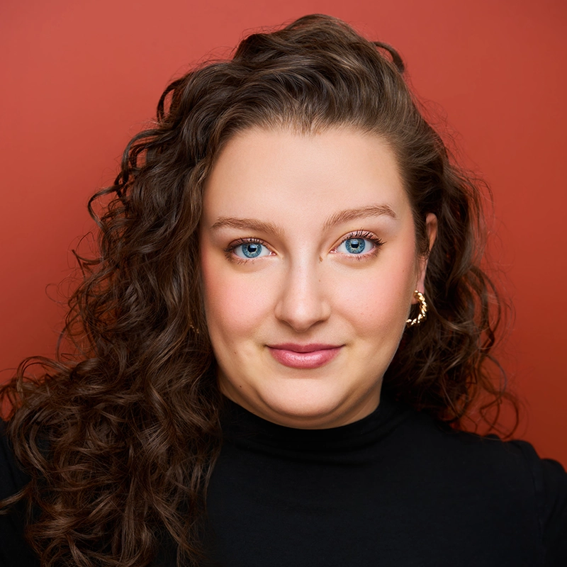 A woman with long, curly brown hair and blue eyes poses against a red background. She is wearing a black turtleneck and gold hoop earrings, and she is giving a soft smile to the camera, evoking the signature portrait style popularized by Spencer Bowman.