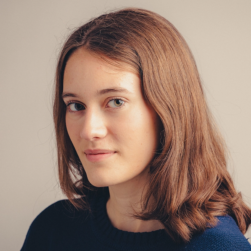 A young woman with fair skin and shoulder-length light brown hair looks at the camera. Gabriela Descartes López is wearing a dark blue sweater and has a slight, subtle smile. The background is plain and softly lit in neutral tones.