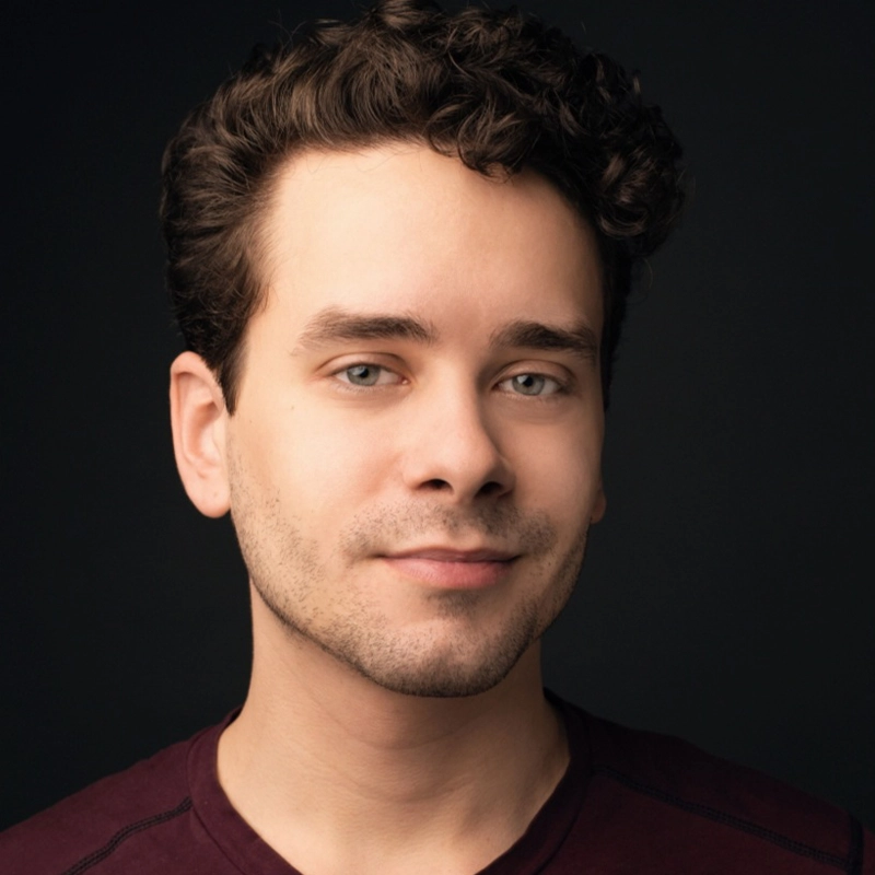 A young man with short, curly brown hair and light skin smiles slightly. He has trimmed facial hair and wears a burgundy shirt. The background is dark, making his face stand out clearly—this is Joe O’Malley.