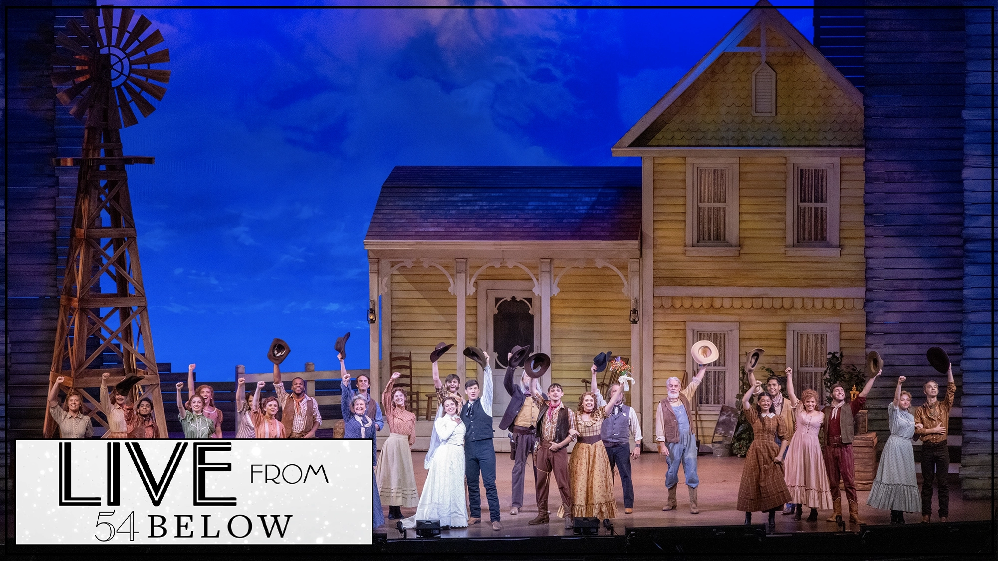 A large cast in colorful period costumes stands on stage in front of a yellow farmhouse set, with many raising hats in triumph. A windmill is left. The scene is lively, set under a blue sky. “LIVE from 54 BELOW” is overlaid in the bottom left corner.