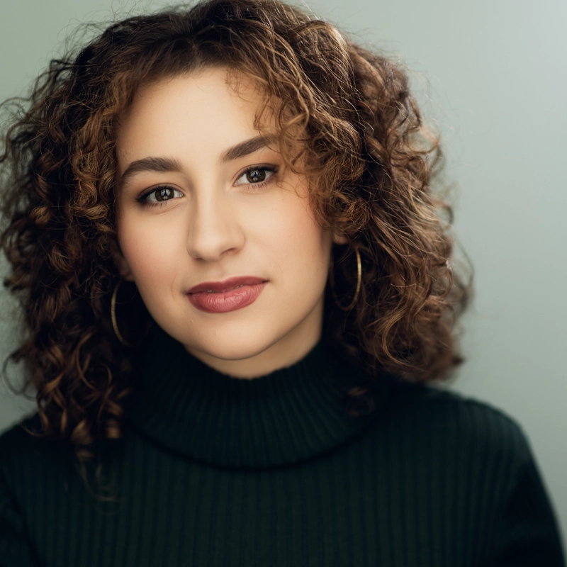 A young woman with light skin and curly brown hair smiles softly, wearing gold hoop earrings and a dark green ribbed turtleneck sweater. The background is a plain, light, cool-toned color. Her makeup is natural and subtle, embodying Olivia Palmer’s effortless style.