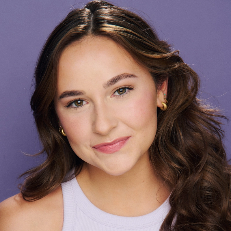 A young woman with long brown wavy hair smiles gently. She wears gold hoop earrings and a sleeveless light lavender top. Her makeup is natural, with pink lipstick and subtle blush. The background is a smooth purple color.