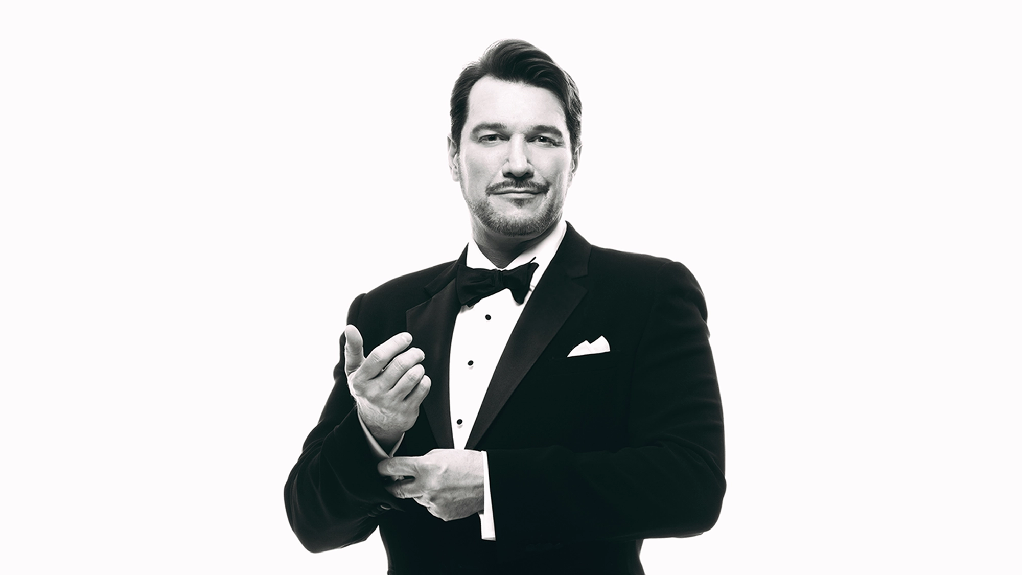 Paulo Szot site image 27 A man in a tuxedo stands against a white background, looking confident with a slight smile, as if ready to take the stage in a musical showdown. He adjusts his cufflinks with one hand while the other hand is raised, and he has short dark hair and light facial hair. The image is in black and white.