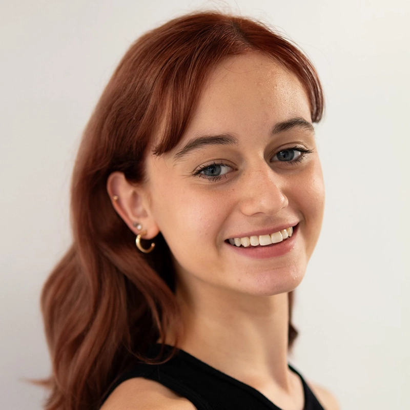 A young woman with long auburn hair smiles at the camera. She has fair skin, blue eyes, and is wearing a black sleeveless top. Small gold hoop earrings and a few ear piercings accent her look. The background is plain and softly lit, drawing focus to Phoebe Kleehammer’s warm expression.