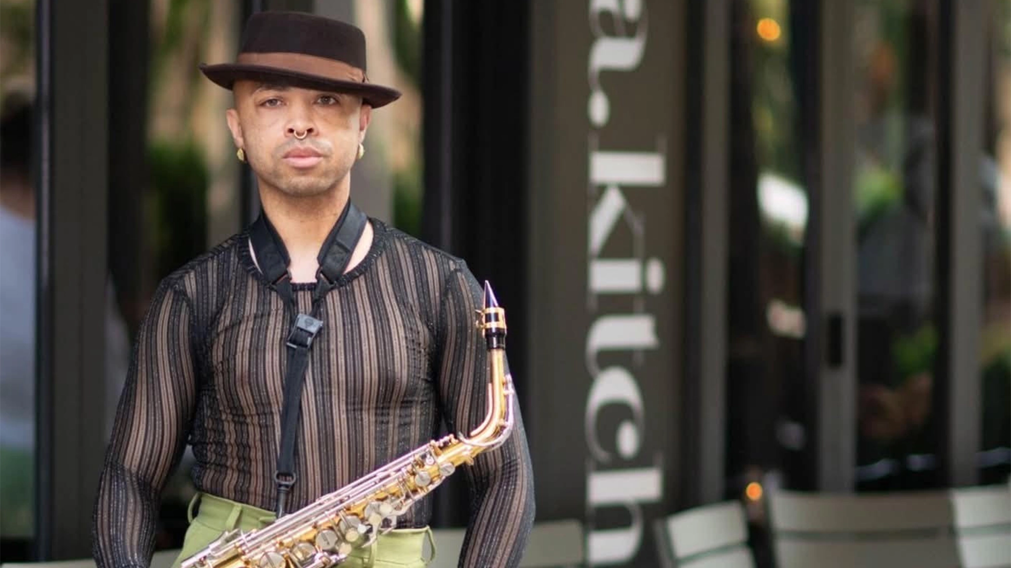 Quinn Piper site image 27 A person wearing a sheer black striped shirt, green pants, and a brown hat stands outside holding a saxophone, as if ready for a concert. The background shows glass doors and the partial text “.kitch” on a sign. The person faces forward with a neutral expression, embodying an effortless cool that says, “Reject Me Already.”.