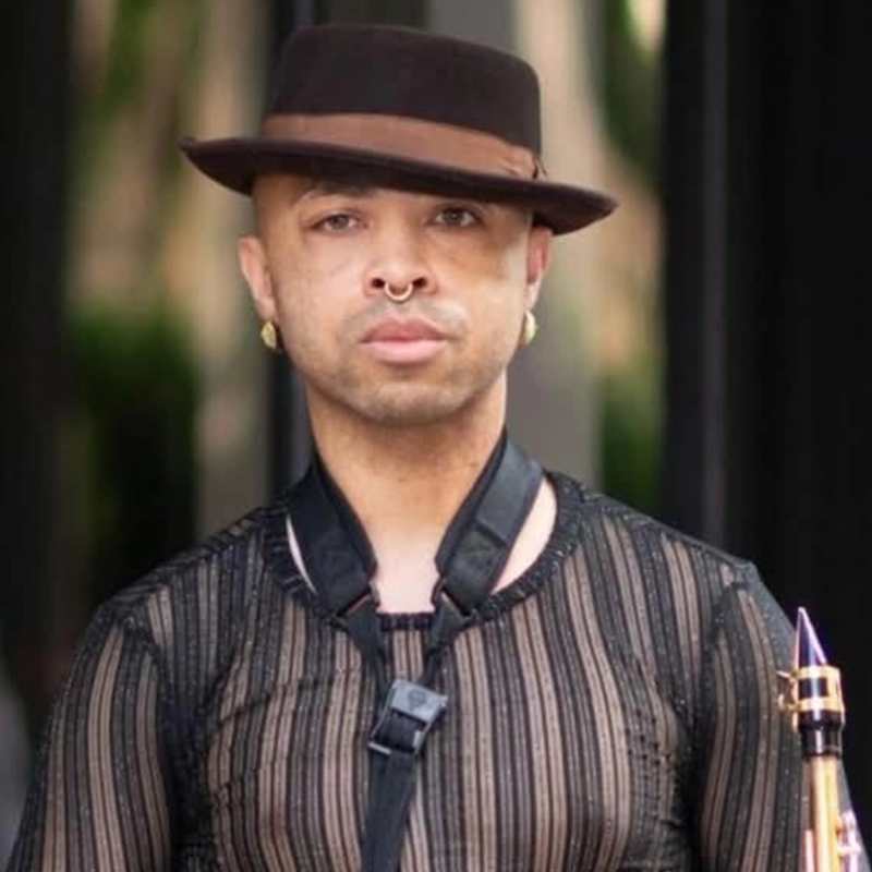 A person with short hair wears a brown fedora hat, gold hoop earrings, and a septum ring. They have a serious expression and wear a sheer, vertically striped black shirt with a saxophone strap over their shoulders. Green foliage is blurred in the background.