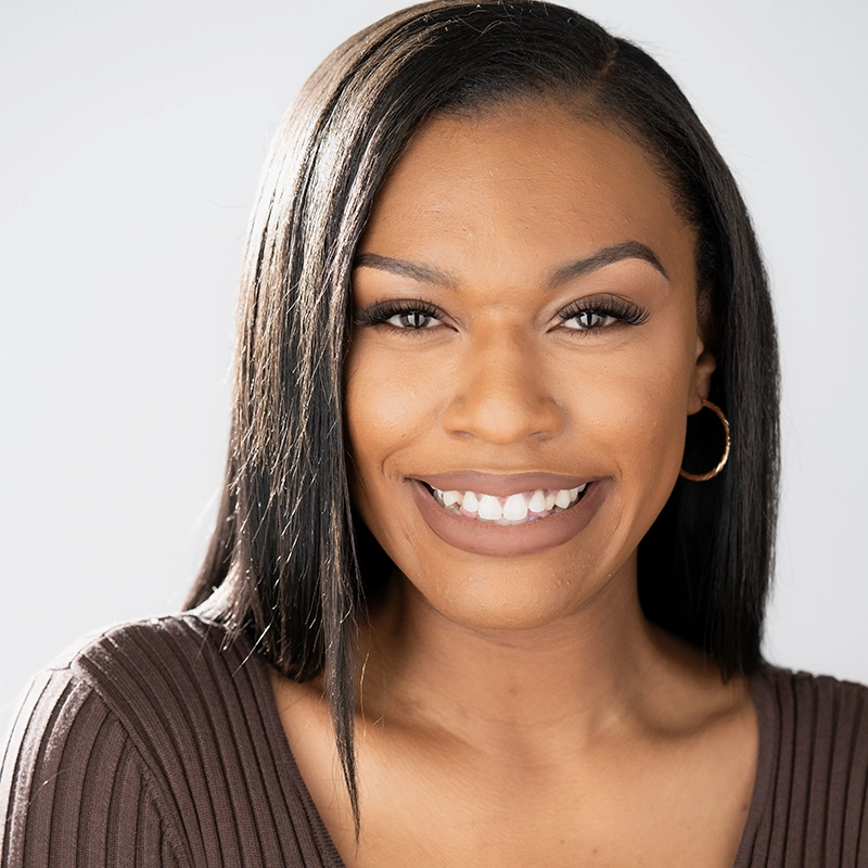 Rayana D. Briggs, a woman with straight, dark hair, smiles at the camera. She wears a brown ribbed top and gold hoop earrings. Her makeup is natural, with neatly shaped eyebrows and soft lipstick. The background is plain and light, highlighting her face.