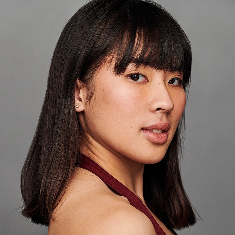 A person with straight, dark brown shoulder-length hair and bangs faces slightly sideways, looking at the camera. They have smooth skin, light makeup, and wear a sleeveless burgundy top. The background is plain gray, giving the portrait a refined and contemporary feel reminiscent of Jonathan Uber’s photographic style.