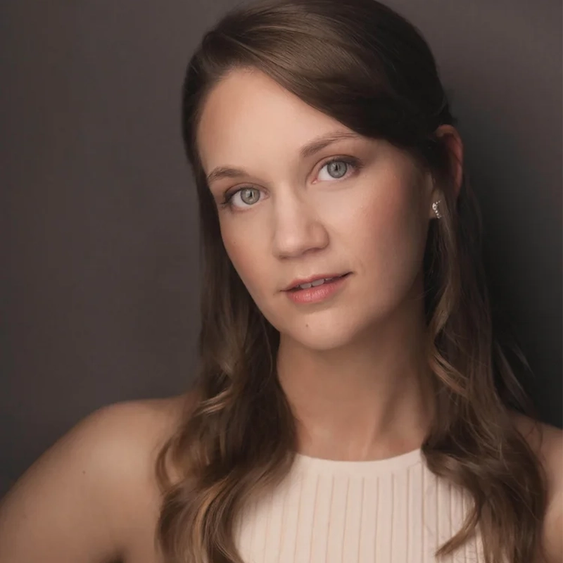 Katherine Riddle, a woman with wavy brown hair and blue eyes, poses against a dark background. She has fair skin, wears subtle makeup, and a sleeveless cream top with vertical pleats. She looks slightly to the side with a gentle, neutral expression.