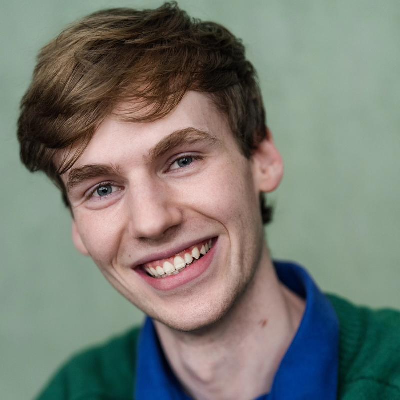 Young adult with light skin and short, light brown hair smiles at the camera. He wears a green sweater over a blue collared shirt. The background is a soft, light green, and the overall mood is cheerful and friendly.